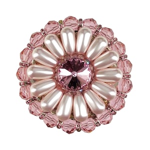 以下が含まれることがあります： 中央に大きなピンク色のクリスタルをあしらった、ピンクと白の花のブローチ。ブローチはフェイクパールとラインストーンでできています。