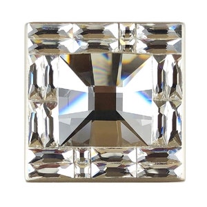 Puede incluir: Un adorno de cristal de forma cuadrada con un centro claro y facetado rodeado por un borde de cristales más pequeños, claros y facetados.