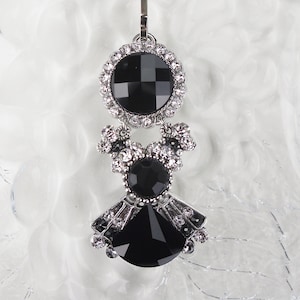 May include: A silver charm with a black geometric design. The charm features a large, round black gem at the top, a smaller black gem in the middle, and a fan-shaped black gem at the bottom, all accented with clear crystals.