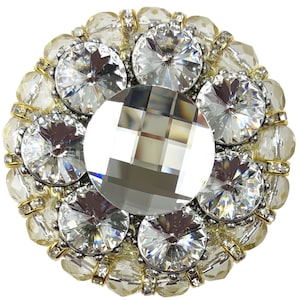 Może przedstawiać: Duża, okrągła, złocona broszka z centralnym, przezroczystym kryształem otoczonym mniejszymi, przezroczystymi kryształami. Broszka jest ozdobiona małymi, przezroczystymi koralikami.