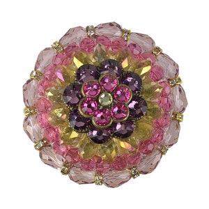 以下が含まれることがあります： ピンク、パープル、ゴールドのジュエリーが飾られた花のブローチ。ブローチはガラスビーズとラインストーンでできています。