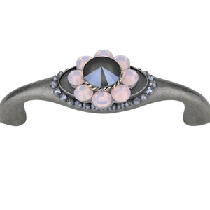 Puede incluir: Un tirador de armario plateado con un diseño decorativo de cristal. El cristal central es negro con un diseño facetado, rodeado por un anillo de cristales rosas más pequeños.