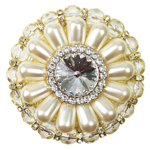 Może przedstawiać: Duży, okrągły broszka z białych pereł z dużym, przezroczystym kryształem w środku. Broszka jest otoczona mniejszymi, przezroczystymi kryształami i złotymi akcentami.