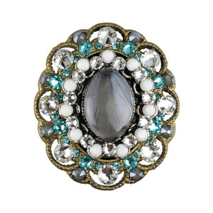 May include: A gray oval gemstone surrounded by a ring of clear and blue rhinestones set in a gold-toned metal frame.