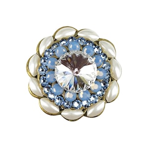 以下が含まれることがあります： 中央に大きな透明なクリスタルがついた、丸い白と青の宝石のブローチです。