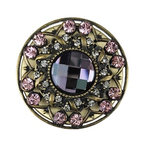 May include: A round, ornate, gold-toned brooch with a large, faceted, purple gemstone surrounded by smaller, pink gemstones.