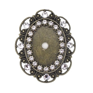 以下が含まれることがあります： 装飾的なデザインと透明なクリスタル製の縁取りが施された、楕円形の金属フレーム。フレームはアンティークゴールドで、中央に小さな穴があります。