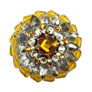 May include: A round brooch featuring a large, square, amber-coloured gemstone surrounded by smaller, clear and yellow gemstones. The brooch is set in silver and has an intricate design.