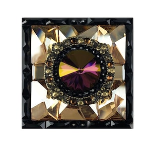 May include: A square-shaped, black framed, decorative piece with a central round, iridescent purple gemstone surrounded by smaller, gold-toned gemstones. The piece is made of glass and has a mosaic-like design.