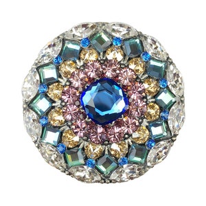May include: A round, silver-toned brooch with a blue, pink, and gold jeweled design. The brooch is surrounded by clear crystals.