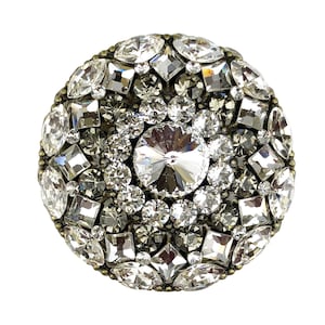 以下が含まれることがあります： 中央に大きな透明なクリスタルがあり、その周りを様々なサイズと形の小さなクリスタルが囲んでいる、丸い銀色のブローチです。