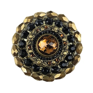 以下が含まれることがあります： 中央に大きな琥珀色のラインストーンが付いた、丸い金色調のボタン。ラインストーンは、小さな黒いラインストーンの円と、大きな金色のビーズの円に囲まれています。