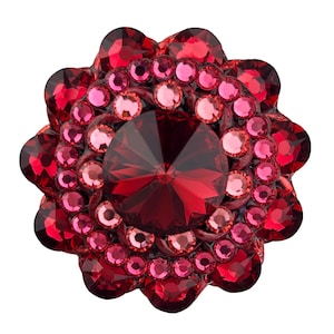 Puede incluir: Una joya redonda y ornamentada, de color rojo y rosa. El centro presenta una gran piedra facetada de color rojo oscuro, rodeada de círculos concéntricos de gemas rosas y rojas más claras. El diseño se asemeja a una flor o a una explosión estelar.