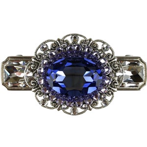 Op de afbeelding: Een zilveren haarspeld met een grote, ovale, donkerblauwe edelsteen omringd door kleinere, heldere en paarse strass steentjes.