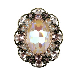 May include: An oval-shaped brooch featuring a large iridescent crystal surrounded by smaller crystals in shades of pink and clear. The brooch is set in an antique gold-toned metal frame.