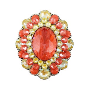 May include: A large, oval-shaped brooch with a central red crystal surrounded by smaller yellow and red crystals.