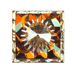 Peut inclure: Une broche carrée dorée avec un grand cristal facetté de couleur ambre au centre. Le cristal est entouré d'un motif en forme d'étoile éclatante argenté.