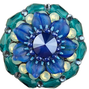 May include: A flower-shaped brooch featuring a central dark blue faceted crystal, surrounded by blue petals. The petals are encircled by pale yellow and teal crystals, and teal geometric shapes.