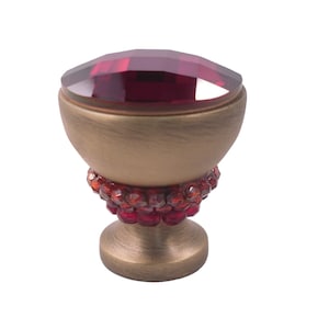 以下が含まれることがあります： 金色調のキャビネットノブ。大きな赤いファセットカットのクリスタルが上部にあり、ベースの周りに小さな赤いクリスタルが並んでいます。