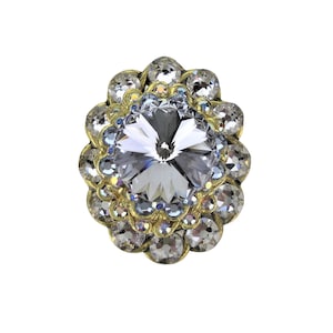 Może przedstawiać: Duży, przezroczysty kryształowy strass otoczony pierścieniem mniejszych, przezroczystych strassów. Strassy są osadzone w złotej oprawie.