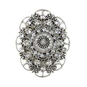 以下が含まれることがあります： 中央に大きなクリアなラインストーンのクラスターがあり、その周りを小さなラインストーンが囲む、円形のデザインの銀色のブローチです。