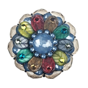 Puede incluir: Un broche redondo y multicolor con un diseño floral. El broche presenta una gran piedra central azul facetada, rodeada de gemas en forma de lágrima en tonos rojos, verdes, marrones y amarillos. Piedras claras y facetadas forman los pétalos exteriores.