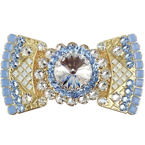 Könnte beinhalten: Eine goldfarbene Brosche in Form einer Schleife mit einem großen, klaren Kristall, der von kleineren blauen Kristallen umgeben ist. Die Brosche ist mit einem geometrischen Muster aus Weiß und Gold verziert.