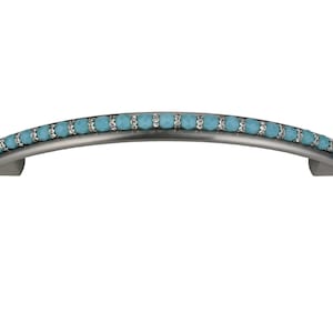 Puede incluir: Un tirador de armario de tono plateado con un diseño curvo. El tirador está acentuado con una fila de cuentas de vidrio azul turquesa.