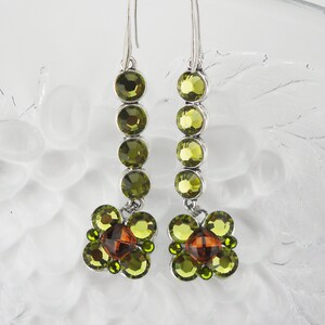 May include: A pair of silver dangle earrings with a cluster of green and brown crystals. The earrings feature a row of five round green crystals followed by a cluster of smaller green and brown crystals.