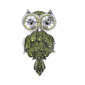 Puede incluir: Un broche de búho verde y plateado con detalles de cristal. Los ojos del búho están hechos de cristales transparentes con un respaldo plateado. El cuerpo del búho está cubierto de cristales verdes.