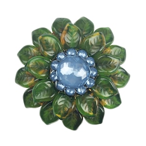 Könnte beinhalten: Eine grüne und blaue Blumenbrosche mit einem großen blauen Strassstein in der Mitte. Die Blume besteht aus mehreren grünen Blättern mit einem goldartigen Schimmer.