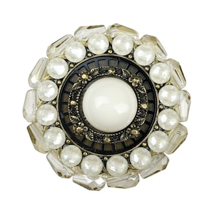 Op de afbeelding: Een ronde, sierlijke broche met een witte middelste steen omringd door heldere en witte kralen. De broche heeft een gouden en zwart metalen frame.