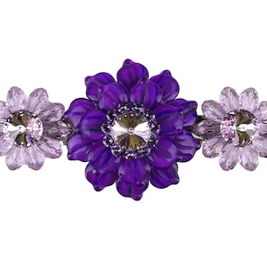 Puede incluir: Una pinza para el pelo con forma de flor morada con tres acentos de flores. La flor central es una flor morada grande con un centro de cristal de roca transparente. Las dos flores más pequeñas son de color morado claro con cristales de roca transparentes en el centro.