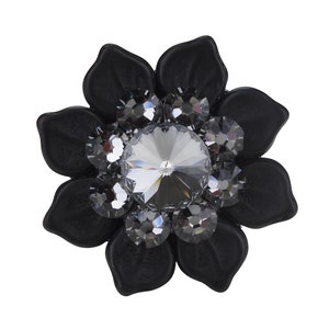 Może przedstawiać: Czarna broszka w kształcie kwiatu z dużym, przezroczystym kryształem w środku i mniejszymi szarymi kryształami wokół.
