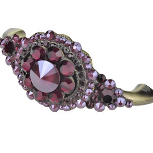 May include: A decorative cabinet pull with an antique brass finish and a large, faceted, burgundy crystal in the center. The crystal is surrounded by smaller, burgundy crystals.