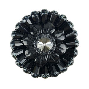Könnte beinhalten: Eine schwarze, blumenförmige Brosche mit einem großen, klaren Strassstein in der Mitte. Die Brosche besteht aus schwarzen Perlen und hat eine glänzende Oberfläche.