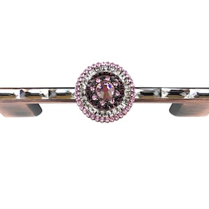 Könnte beinhalten: Ein dekorativer Schrankgriff mit einem zentralen kreisförmigen Design mit einem großen rosa Kristall, umgeben von kleineren rosa und klaren Kristallen. Der Griff hat eine Bronzefinish und rechteckige klare Kristalle.