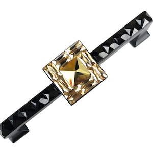 Könnte beinhalten: Ein schwarzer Schrankgriff mit facettiertem Design. In der Mitte befindet sich ein quadratischer, goldfarbener Kristall. Der Griff ist etwa 15 cm lang.