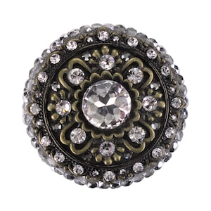 以下が含まれることがあります： 中央に大きな透明なクリスタルがついた、丸い装飾的な銀色のボタン。ボタンは、小さなクリスタルと装飾的な花柄のデザインで囲まれています。