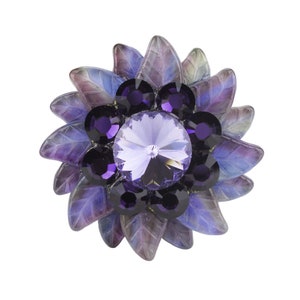 Könnte beinhalten: Eine violette, blumenförmige Brosche mit einem großen, klaren Kristall in der Mitte und kleineren violetten Kristallen darum herum.