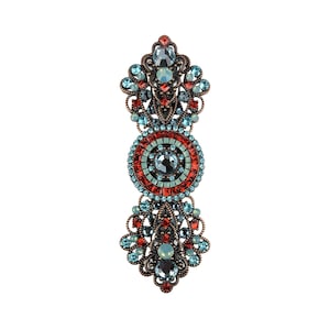 Peut inclure: Une broche décorative avec un motif circulaire au centre, entouré de cristaux rouges et bleus. La broche a un motif floral en haut et en bas, également orné de cristaux rouges et bleus.