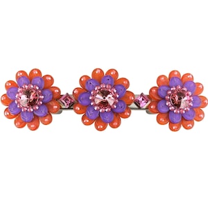 Puede incluir: Una pinza para el pelo con tres abalorios en forma de flor naranja y morada con pedrería rosa en el centro.