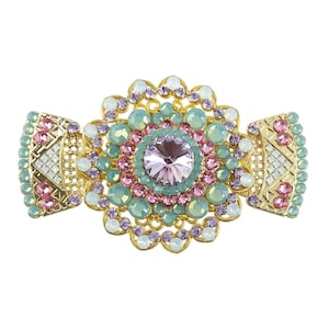 以下が含まれることがあります： 多色の宝石で飾られた大きな中心部を持つ金色のブローチ。中心部には、薄い紫色の宝石が、より小さな緑色、ピンク色、白色の宝石で囲まれています。ブローチは、側面に幾何学模様が施されています。