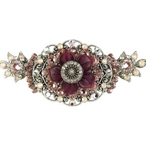 Op de afbeelding: Een zilverkleurige haarspeld met een grote, donkerpaarse bloem in het midden. De bloem is omgeven door kleinere roze en heldere strasssteentjes. De speld heeft een delicaat, sierlijk ontwerp.