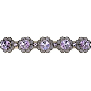 Puede incluir: Una pinza para el pelo de plata con cinco adornos en forma de flor. Cada flor tiene un gran strass morado iridiscente claro en el centro y strass claros más pequeños alrededor del borde.