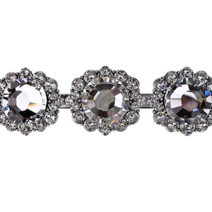 Może przedstawiać: Trzy srebrne, okrągłe broszki z przezroczystymi kryształami w środku i mniejszymi, przezroczystymi kryształami otaczającymi centralny kamień.