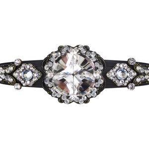 Può includere: Un fermaglio per capelli nero con un grande cristallo trasparente circondato da cristalli più piccoli. Il fermaglio ha un design decorativo e ornato.