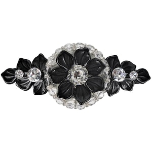 Może przedstawiać: Czarna broszka w kształcie kwiatu z przezroczystymi cyrkoniami. Broszka ma duży centralny kwiat z mniejszymi kwiatami po obu stronach.