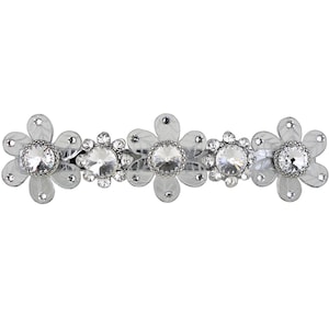 Puede incluir: Una pinza para el pelo plateada con tres flores de plástico transparente, cada una con un gran strass transparente en el centro.