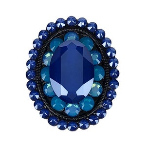 May include: An oval-shaped brooch featuring a large, faceted, dark blue gemstone surrounded by a ring of smaller, light blue gemstones.